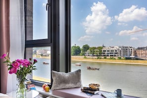 Una habitación con un ventanal que da a un río, un florero, un cojín, libros y una taza sobre una mesa.