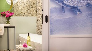 A bathroom with a mosaic tile wall, a mirror, a sink with a towel, a vase with flowers, and a wine bottle on a tray.