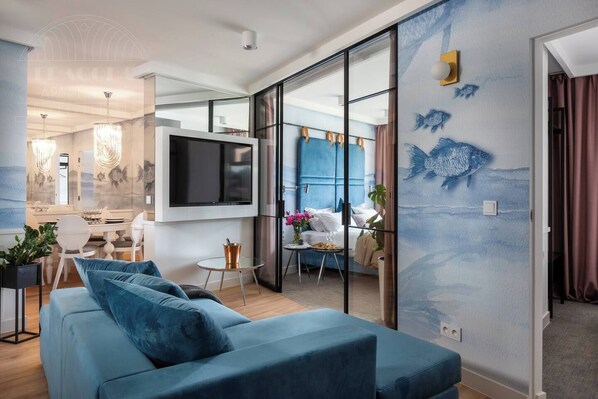 A modern living room with a blue sofa, a flat-screen TV, and a dining area with a table and chairs. - Apartment Vistula 3-bed with a balcony and bathtub (Kraków - Vistula View)