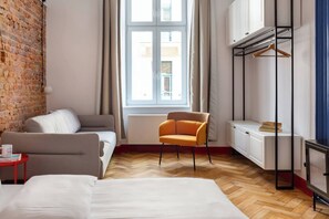 A hotel room with a brick wall, a sofa, a chair, a bed, a radiator, a wardrobe, and a window with curtains. - The Geometric 3-bedroom apartment (Kraków – Old Town)