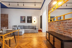 Un salon moderne avec un parquet en bois à chevrons, un canapé gris, un miroir à cadre jaune et un mur d’accent en briques.