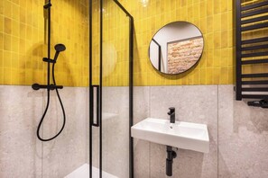 Une salle de bain avec des murs recouverts de carreaux jaunes, une douche en verre, un lavabo blanc et un miroir rond qui reflète un mur en briques.
