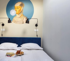 A hotel room with a bed, pillows, a wall-mounted lamp, a painting, and a plate with food.