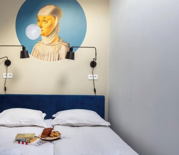 A hotel room with a bed, pillows, a wall-mounted lamp, a power outlet, a plate with food, and a book.