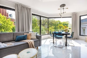 A modern living room with a sofa, dining table, chairs, and large windows offering a view of greenery and buildings. - Apartment Modern Spacious (Kraków – Modern View of Planty Park)