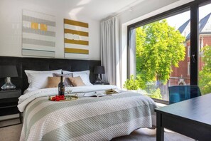 A modern bedroom with a large window, a bed with a white and gray bedspread, a bedside table with a lamp, and a view of a tree and building outside. - Apartment Modern Spacious (Kraków – Modern View of Planty Park)