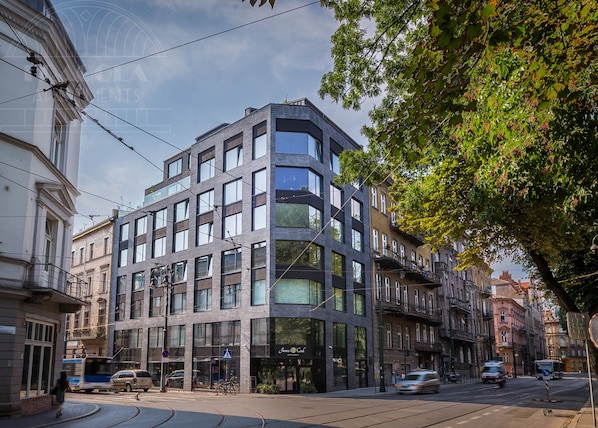 A modern multi-story building with large windows, a restaurant on the ground floor, and a tram line nearby. - Apartment Modern Spacious (Kraków – Modern View of Planty Park)