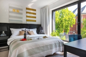 A modern bedroom with a large window, a bed with a white and gray bedspread, a bedside table with a lamp, and a view of a tree and building outside. - Apartment Modern Spacious (Kraków – Modern View of Planty Park)