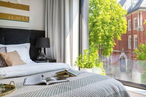A hotel room with a bed, a nightstand, a lamp, a window with a view of a red brick building and greenery. - Apartment Modern Spacious (Kraków – Modern View of Planty Park)