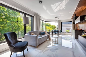 A modern living room with a sofa, a dining table, and chairs, large windows with a view of trees, and a kitchen area with cabinets and a countertop. - Apartment Modern Spacious (Kraków – Modern View of Planty Park)