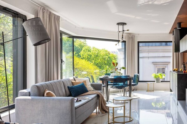 A modern living room with a sofa, a dining table, and chairs. There is a large window providing a view of greenery outside.