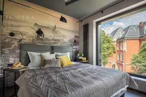 Un dormitorio moderno con un mural de paisaje urbano, una cama grande con ropa de cama estampada y vistas a un edificio de ladrillo rojo.