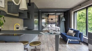 A modern interior with a kitchen island, bar stools, a blue sofa, and a large window offering a view of greenery. - Apartment Modern Industrial (Kraków – Modern View of Planty Park)
