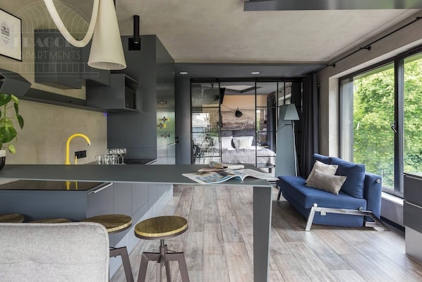 Interior moderno com ilha de cozinha, banquetas, um sofá azul e uma grande janela com vista para o verde.