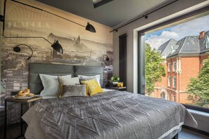 Un dormitorio moderno con un mural de paisaje urbano, una cama grande con ropa de cama estampada y vistas a un edificio de ladrillo rojo.