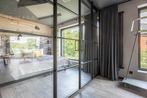 Une chambre moderne avec une cloison en verre, un lit avec des oreillers, un bureau avec une lampe et une vue sur la verdure à l’extérieur.
