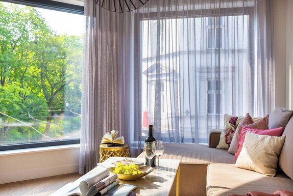 A room with a large window, a sofa, a table with a bottle and glasses, and a bowl of grapes. - Apartment Modern Marble (Kraków – Modern View of Planty Park)