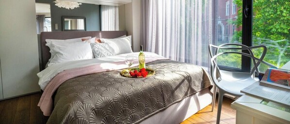 A hotel room with a bed, a dining table with a chair, a window with a view of greenery, and a mirror on the wall. - Apartment Modern Marble (Kraków – Modern View of Planty Park)