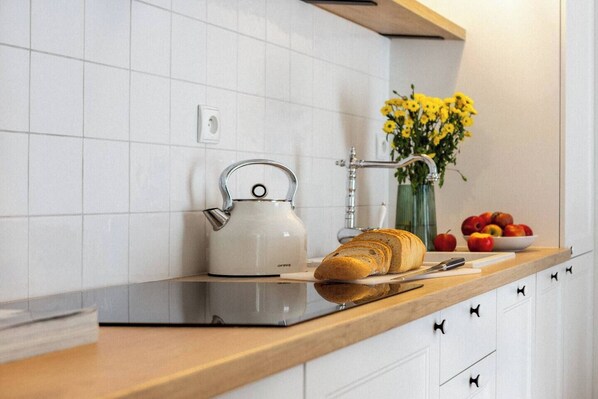 A kitchen with white tiles, a kettle, a loaf of bread, a vase with yellow flowers, and a bowl of fruit. - Canvas House Apartament Oatmeal (Kraków)
