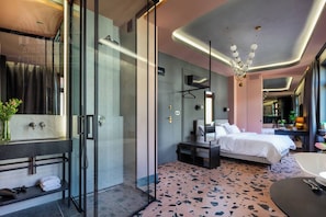 Una habitación de hotel moderna con cabina de ducha de vidrio, una cama grande, una mesita de noche y una pequeña zona de estar.