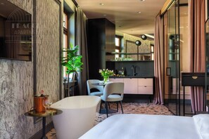 Une chambre d’hôtel moderne dotée d’un grand lit, d’un coin salon avec une table et des chaises, d’un petit coin cuisine et d’une grande fenêtre avec des rideaux.