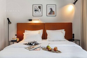 A neatly made bed with white linens, two pillows, a magazine, and three apples on the bed. There are two framed pictures on the wall above the bed, a nightstand with a lamp, and a small round table with a book and a mug. - Canvas House Studio Pretzel with a Balcony (Kraków)
