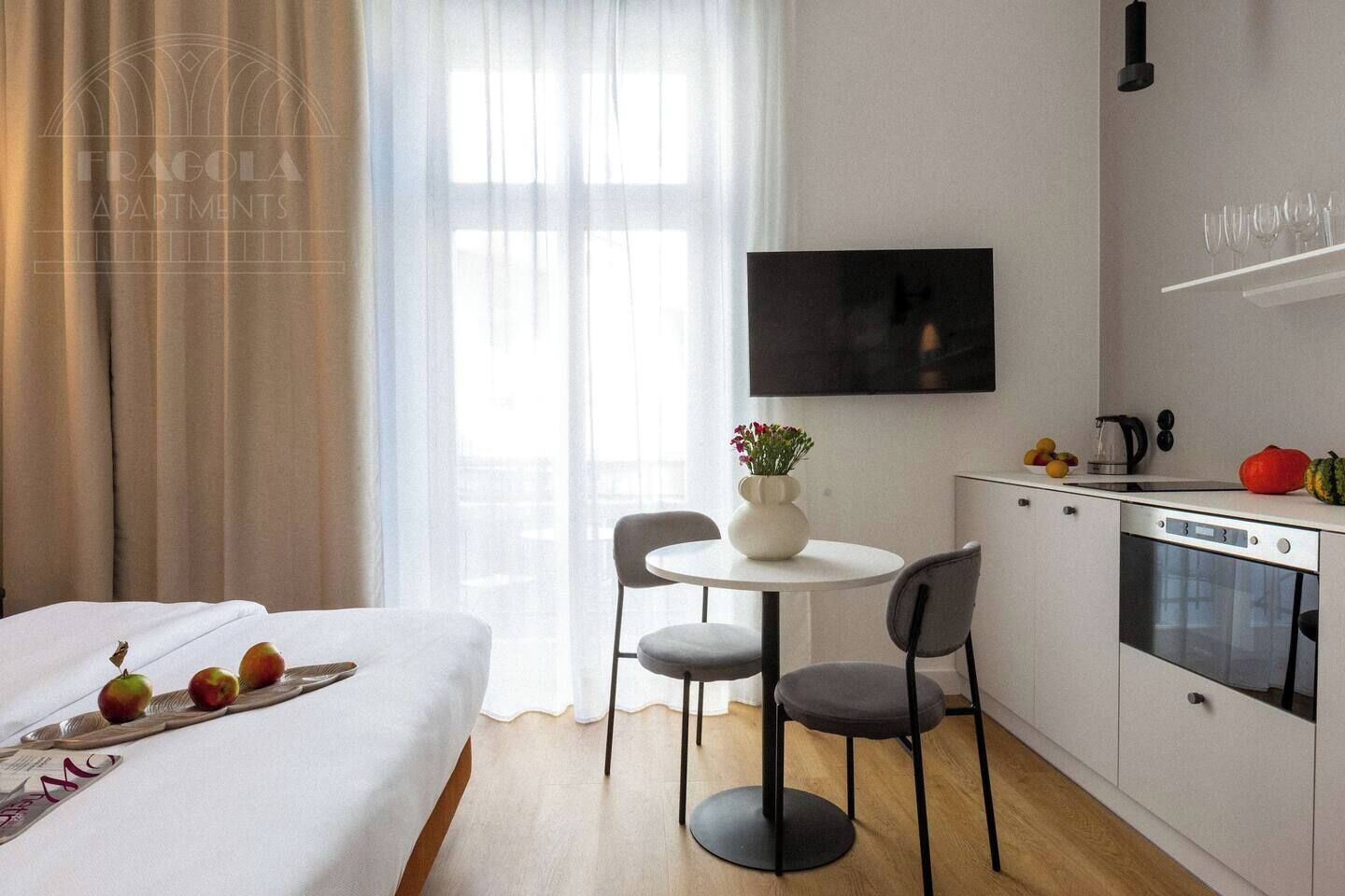 A hotel room with a bed, a dining table with chairs, a kitchen area with a TV, and a window with curtains.