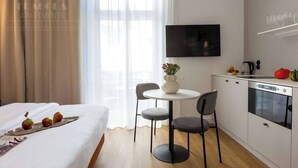 A hotel room with a bed, a dining table with chairs, a kitchen area with a TV, and a window with curtains.