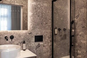 A bathroom with a marble wall, a sink, a glass shower enclosure, and a window with curtains.