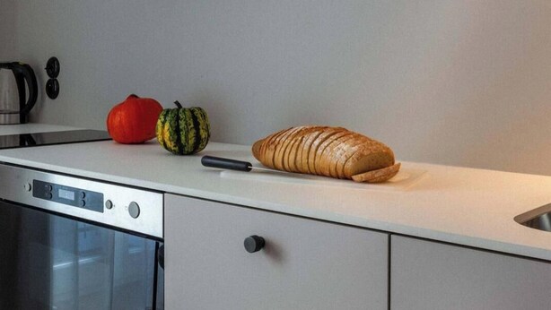 A kitchen with a white countertop, a loaf of sliced bread, a red bell pepper, and a small green squash.