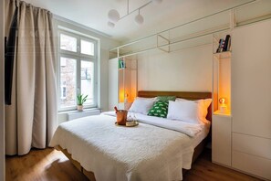 A bedroom with a bed, a window with curtains, a bedside table with a plant, and a wall-mounted shelf with books. - Canvas House Apartament Retro (Kraków)