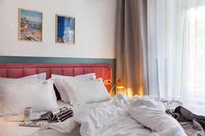 Une chambre d’hôtel avec une tête de lit recouverte de tissu rouge, du linge de lit blanc, un lit avec un magazine et une tasse, et deux tableaux encadrés accrochés au mur.