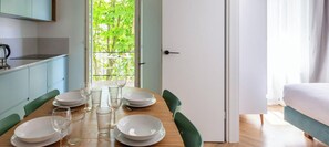 Une table à manger dressée avec des assiettes et des verres, un coin cuisine avec des placards et une porte donnant sur une pièce où se trouve un lit.