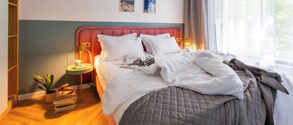 Un lit bien fait, avec des oreillers et une couverture, une table de chevet avec une lampe et une tasse, une petite plante et une fenêtre avec des rideaux.