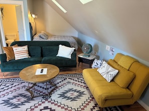 A living room with a green sofa, a yellow armchair, a round coffee table, a patterned rug, a small side table with a fan, and a bed with patterned bedding.