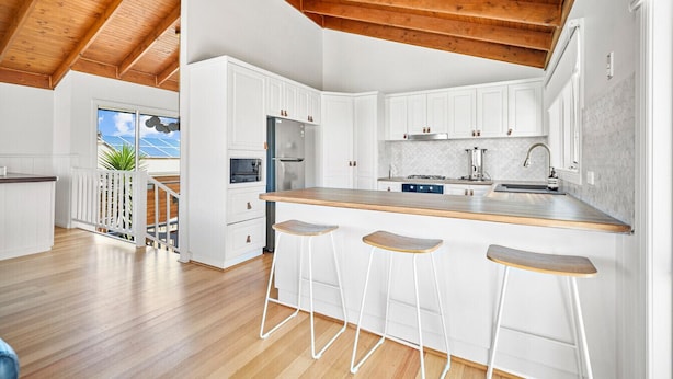 A modern kitchen with white cabinets, a wooden ceiling, and a bar area with stools. - The Palm House 200 meters to the beach! (Torquay)