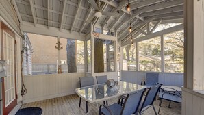 A covered outdoor dining area with a glass table and chairs, wooden deck flooring, and a view of the surrounding landscape.