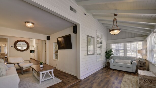A spacious living area with a wooden floor, white walls, and a ceiling with exposed beams. There is a television mounted on the wall, a sofa, a coffee table, and a lamp.
