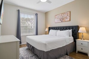 Un dormitorio con cama, mesitas de noche, una ventana con cortinas, un ventilador de techo y una obra de arte colgada en la pared.