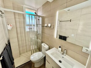 Ein modernes Badezimmer mit einer gläsernen Duschkabine, Toilette, Waschbecken mit Spiegel und einem Fenster über der Dusche.