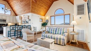 Un interior acogedor con un techo alto de madera, una zona de comedor con sillas de mimbre, un sofá a rayas y una cocina con armarios blancos.