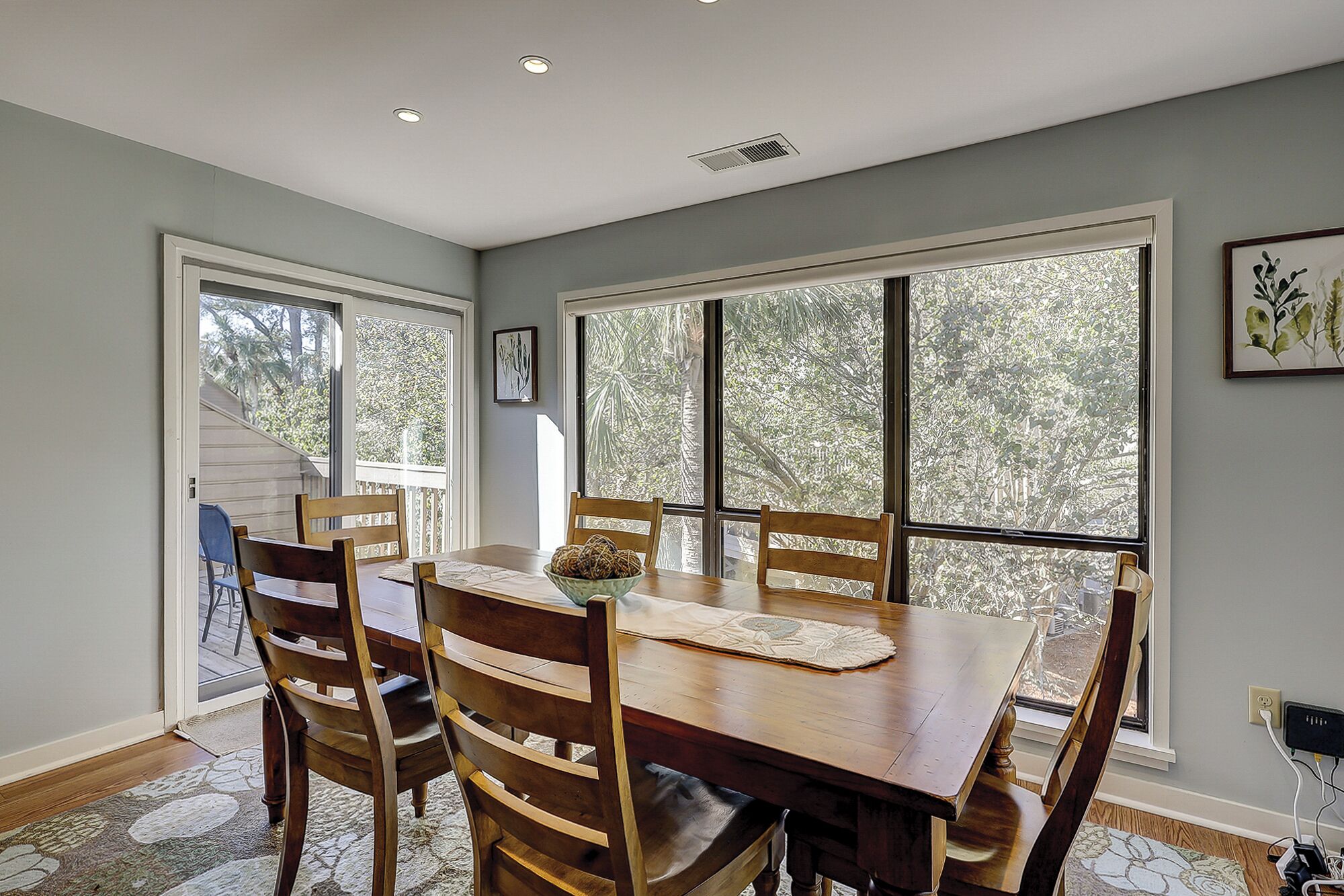 Comedor con mesa y sillas de madera, ventanas amplias con vistas a árboles y un respiradero en el techo.