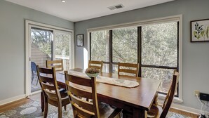 Ein Esszimmer mit Holztisch und -stühlen, großen Fenstern mit Blick auf Bäume und einem Deckenlüfter.