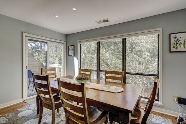A dining room with a wooden table and chairs, large windows with a view of trees, and a ceiling vent.