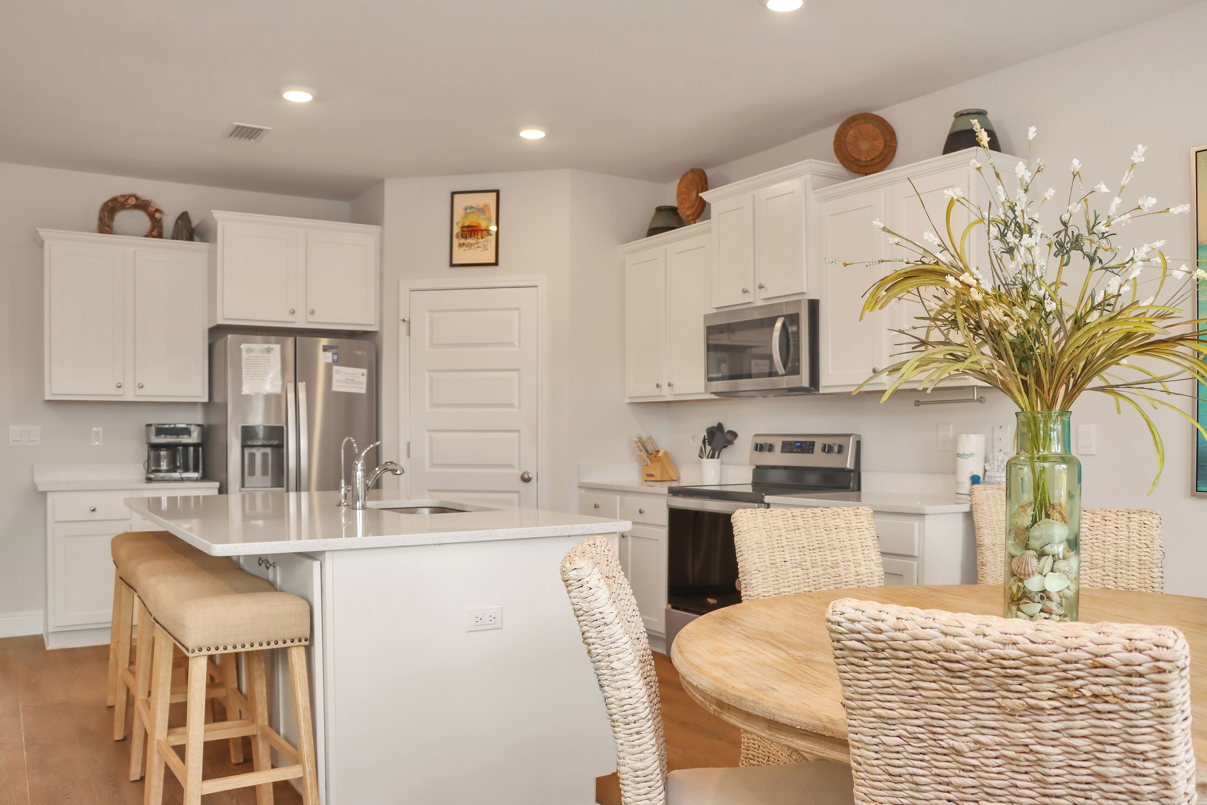 A modern kitchen with white cabinetry, a central island with stools, and a dining area with wicker chairs.