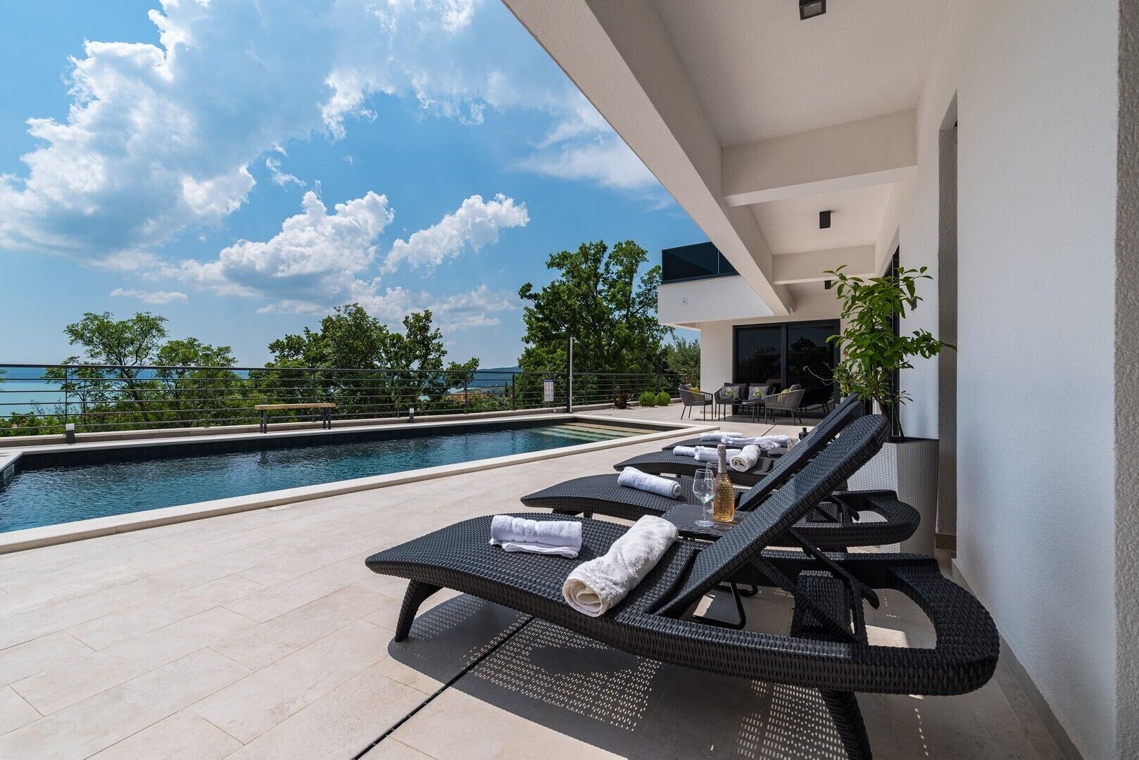 A modern outdoor pool area with lounge chairs, a bench, and a view of the sea.