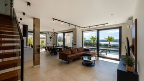 A spacious living area with a large window overlooking a pool and ocean view, a dining area with a table and chairs, and a staircase leading to an upper level. - Luxury Villa Spectrum pool 32m2 sauna, family, sea view, bbq (Posedarje)