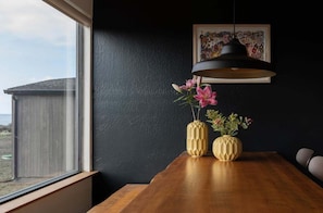 A dining area with a wooden table, two geometric vases with flowers, a black pendant light, and a framed artwork on the wall. - Moorings | Ocean Views | Hot Tub | Dog Friendly (The Sea Ranch)