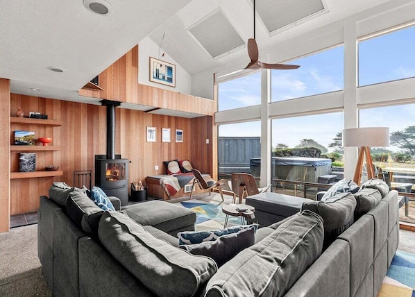 A modern living room with a large grey sectional sofa, a fireplace, wooden panel walls, and large windows offering a view of the outdoors. - Moorings | Ocean Views | Hot Tub | Dog Friendly (The Sea Ranch)