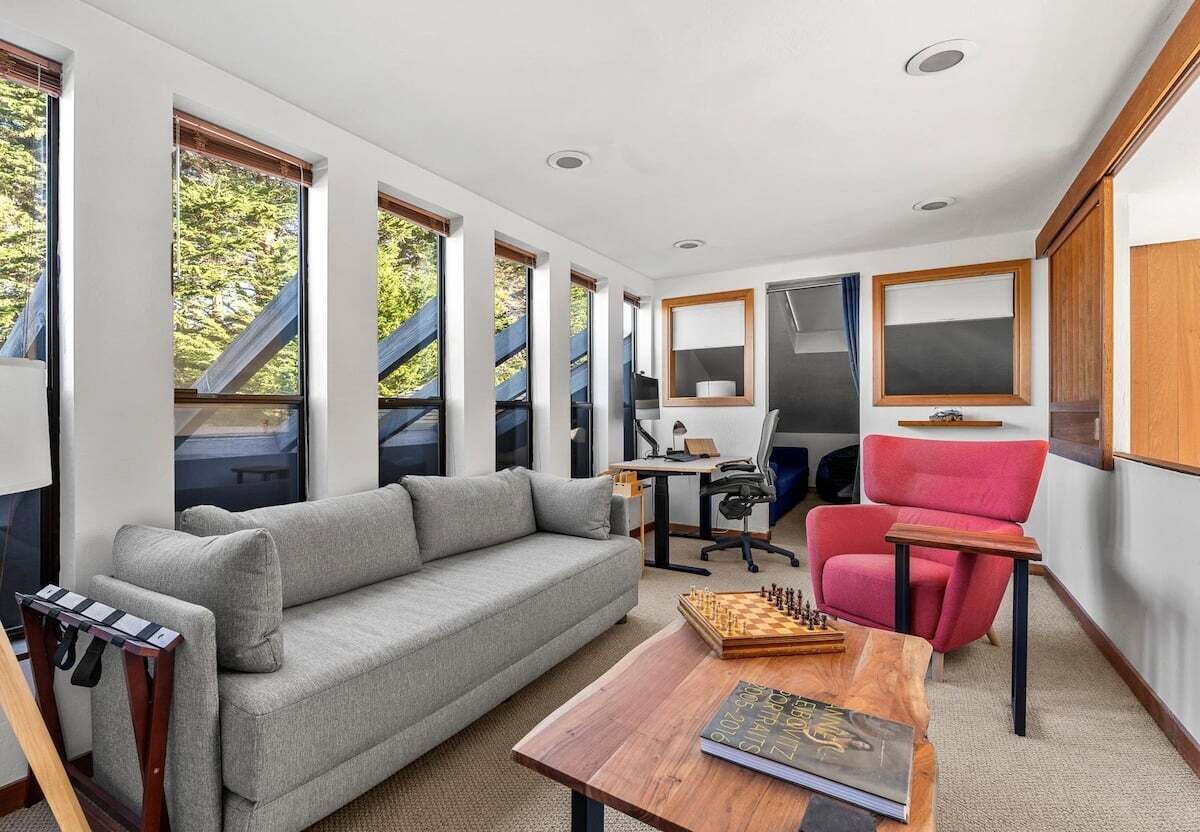 A modern interior with a grey sofa, a red chair, a wooden coffee table with a chessboard, and large windows with a view of trees.